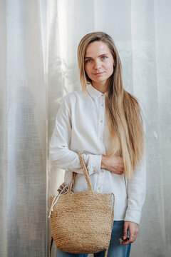 Modest Woman With A Subtle Smile In A White Sunlit Room Posing For A Photo In Front Of A Curtain, Holding Wicker Bag.