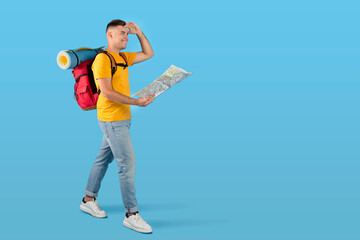 Happy millennial tourist with tourist gears and map looking ahead while walking on blue studio background, copy space