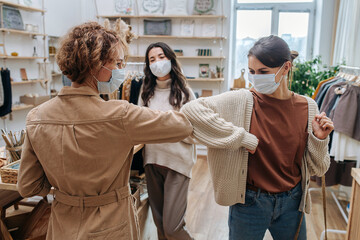 Two curious young women in an ecological shop greeting each other, touching elbows. Third woman behind them. All wearing medical masks.