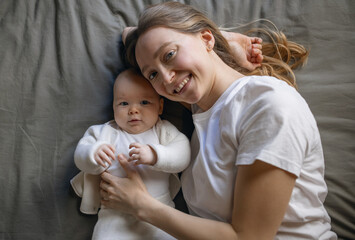 Mom with her baby lies and looks at the camera from the top. Beautiful young woman with her baby. Mother and daughter. Happy family.