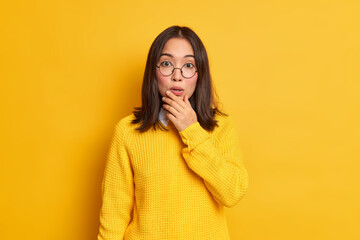 Impressed beautiful Asian woman holds chin looks stunned at camera hears something shocking wears round spectacles casual sweater isolated over yellow background. People and reaction concept