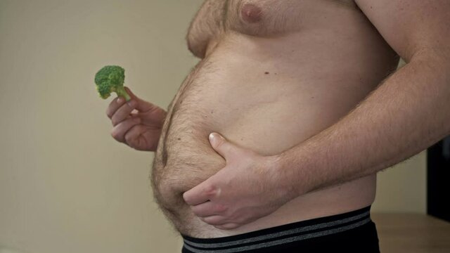 Fat man shows the folds of excess fat on his stomach with one hand and holds broccoli with the other. Problem with being overweight and healthy eating.