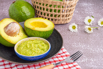 Fototapeta na wymiar Organic avocado with seed, avocado halves, and fresh guacamole in a bowl on a black plate with cloth and cutlery on a gray stone background. Top view. Space for text. Concept of healthy fruit