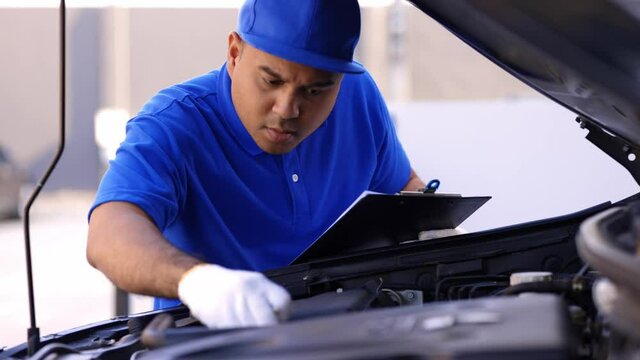A young Asian auto mechanic opens the bonnet. To check for engine damage And perform professional maintenance. He wearing blue uniform.
