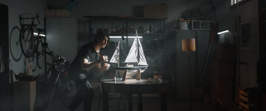 WIDE Caucasian teenager boy making finishing paint touches on boat model project inside his home garage. Shot with 2x anamorphic lens
