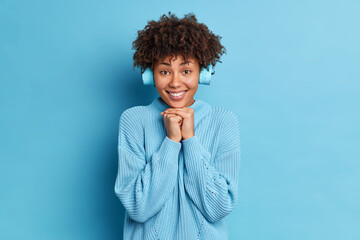 Pretty African American woman keeps hands under chin smiles pleasantly listens audio track via stereo headphones dressed in knitted jumper isolated over blue background. People and lifestyle concept
