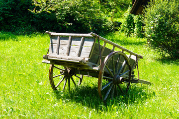 Historic, weathered wooden two-wheeled cart standing on a green meadow surrounded by some bushes.