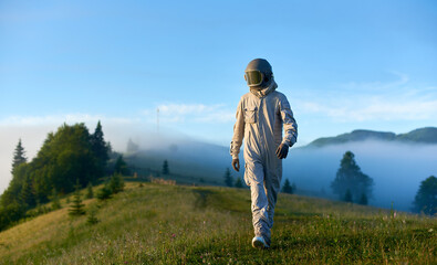 Spaceman wearing white space suit and helmet walking alone sunny green mountain glade in the...