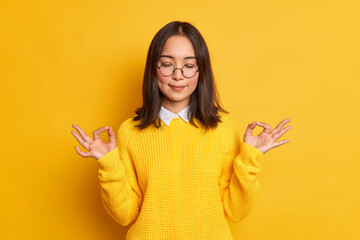Studio shot of of calm young Asian woman shows zen or okay sign meditates in good mood stands with...