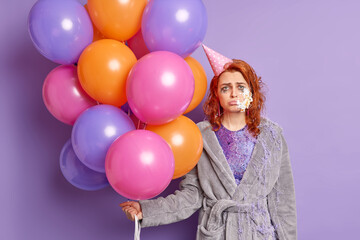 Fototapeta na wymiar Disappointed redhead woman sad to celebrate birthday during quarantine alone has cream on cheek leaked makeup wears domestic robe holds colorful balloons. Upset lonely foxy girl at celebration
