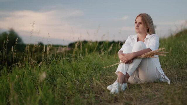 A woman in white clothes rests on a green hill on a warm summer evening