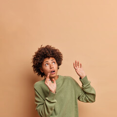 Fototapeta na wymiar Frightened African American woman keeps arms raised tries to stop something falling above concentrated upwards being afraid wears casual jumper poses against beige background copy space area