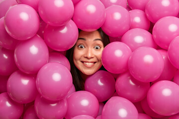 Fototapeta na wymiar Positive cheerful pretty Asian woman looks with great surprisement sticks out head from small inflated airballoons has fun on party expresses happiness celebrates holidays. Festive decorations