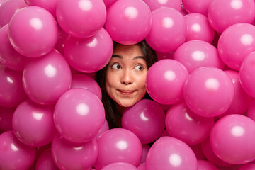 Fototapeta na wymiar Close up shot of brunette Asian woman concentrated aside with interest surrounded by pink helium balloons prepares for party has festive mood. People decoration and special occasion concept.