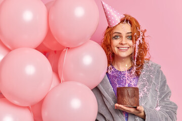 Horizontal shot of happy ginger girl glad after festal occasion has spoiled makeup holds air balloons wears party hat poses with birthday cake poses indoor. Anniversary celebration decoration concept