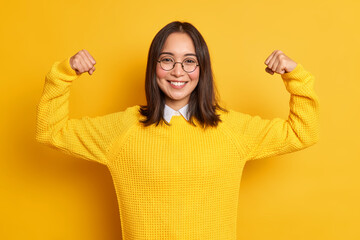 Studio shot of positive young Asian woman raises arms shows muscles pretends to be very strong and...
