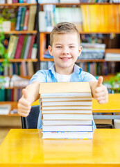 The boy with a smile laid his head on a stack of books in the library and shows class with thumbs of both hands