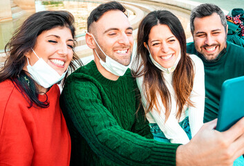 Millennial friends taking selfies smiling by lowering protective face masks - New normal friendship concept with young people having fun together.