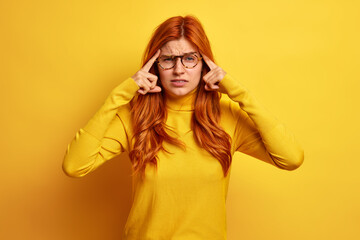 Unhappy redhead woman keeps index fingers on temple has unbearable pain in temples suffers severe migraine wears spectacles and turtleneck isolated over yellow background tries to concentrate