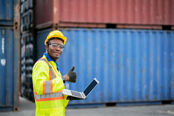 Portrait of African happy worker in protective safety jumpsuit uniform  with hardhat and use laptop computer at cargo container shipping warehouse. transportation import,export logistic industrial