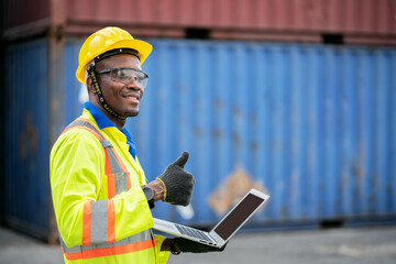 Portrait of African happy worker in protective safety jumpsuit uniform  with hardhat and use laptop computer at cargo container shipping warehouse. transportation import,export logistic industrial