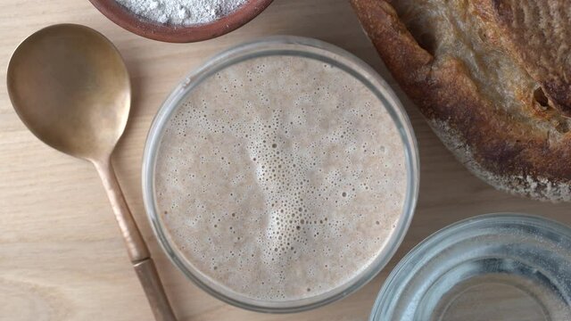 Yeast-free sourdough bread, flour, water and glass jar with dough leaven on the table. Preparing yeast dough for bread, buns, pastries or pizza, close up, top view, revolves