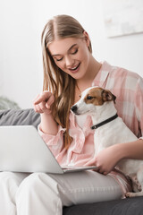 happy woman sitting on couch with jack russell terrier and pointing with finger at laptop