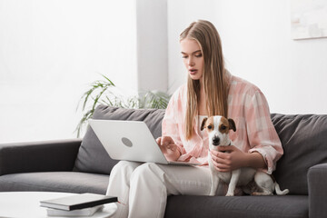 young woman sitting on couch with jack russell terrier and using laptop