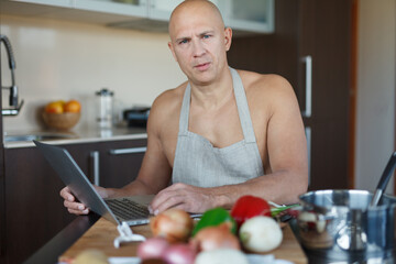 A man with a naked torso and an apron sits at a table in the kitchen and selects a recipe from a laptop.