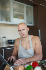 A man with a naked torso and an apron sits at a table in the kitchen and selects a recipe from a laptop.