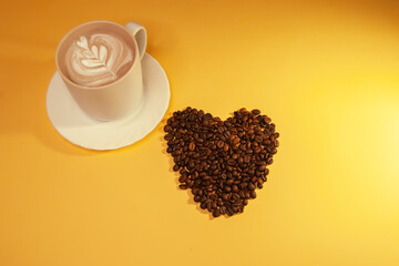Hot coffee with whipped cream and foam in a beige cup and saucer next to a heart of roasted coffee beans on a yellow background.
