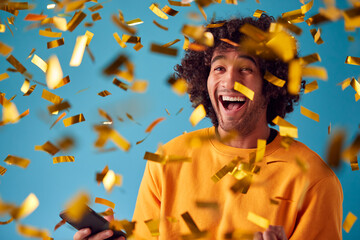 Celebrating Young Man With Mobile Phone Winning Prize And Showered With Gold Confetti In Studio - obrazy, fototapety, plakaty