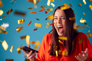 Fototapeta Celebrating Young Woman With Mobile Phone Winning Prize And Showered With Gold Confetti In Studio obraz