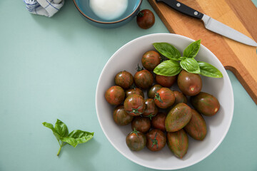 Gestreifte bunte Tiger Tomaten in Schale mit Mozzarella und Basilikum mit Geschirrtuch, Messer, Holz Brett auf Hintergrund Mint Grün