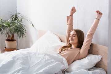 awake and joyful woman in satin pajamas stretching in bed