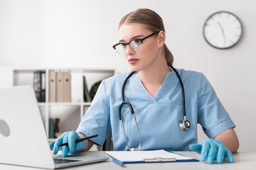 young veterinarian in glasses and latex gloves using laptop near clipboard on table
