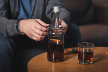 cropped view of man, handcuffed to bottle of whiskey, sitting at home alone