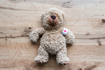 Ein brauner Teddybär mit einem Pflaster und einem Herz auf dem Arm liegt auf einem Holz Tisch. Draufsicht, Impfung, Symbol.