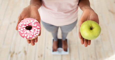 Healthy vs unhealthy food. Black woman holding apple and donut on scales, making choice of her...