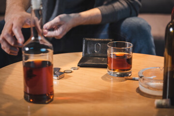 cropped view of alcohol-addicted man taking bottle of whiskey from table near empty wallet, blurred foreground