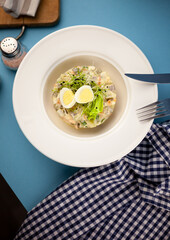 Traditional salad Olivier. Christmas and New year food. White plat with fork and knife on blue background with copy space. Top view, flat lay. Microgreen, quail eggs. Serving at a restaurant.