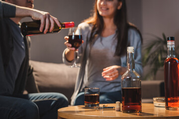 cropped view of husband pouring wine into glass of wife near bottles of alcohol on table, blurred background
