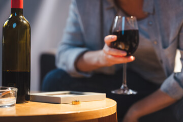 bottle of alcohol, photo frame and wedding ring near woman holding glass of red wine on blurred background