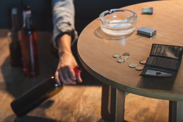 partial view of female hand with bottle of alcohol near coins and empty wallet on table, blurred background