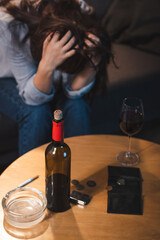 alcohol-addicted woman sitting with bowed head near bottle of red wine, coins and empty wallet, blurred background