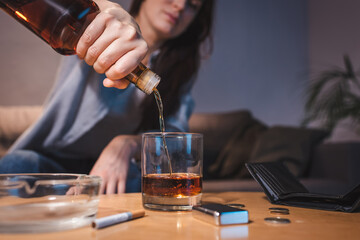 cropped view of woman pouring whiskey into glass near ashtray, coins and empty wallet, blurred background
