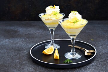 Portion creamy dessert with lemon curd, mascarpone cream and biscuit, garnished with whipped cream and crushed pistachios, in triangular glasses on a black concrete background. Dessert recipes.