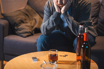Drunk man sitting near table with glass and bottles of alcohol at home
