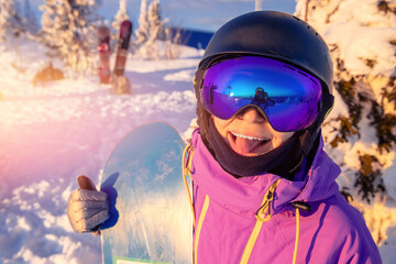 Young happy girl snowboarder holds snowboard on background of sunset and ski resort