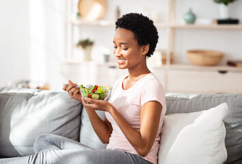 Balanced nutrition concept. Lovely black lady eating delicious vegetable salad on cozy couch at home